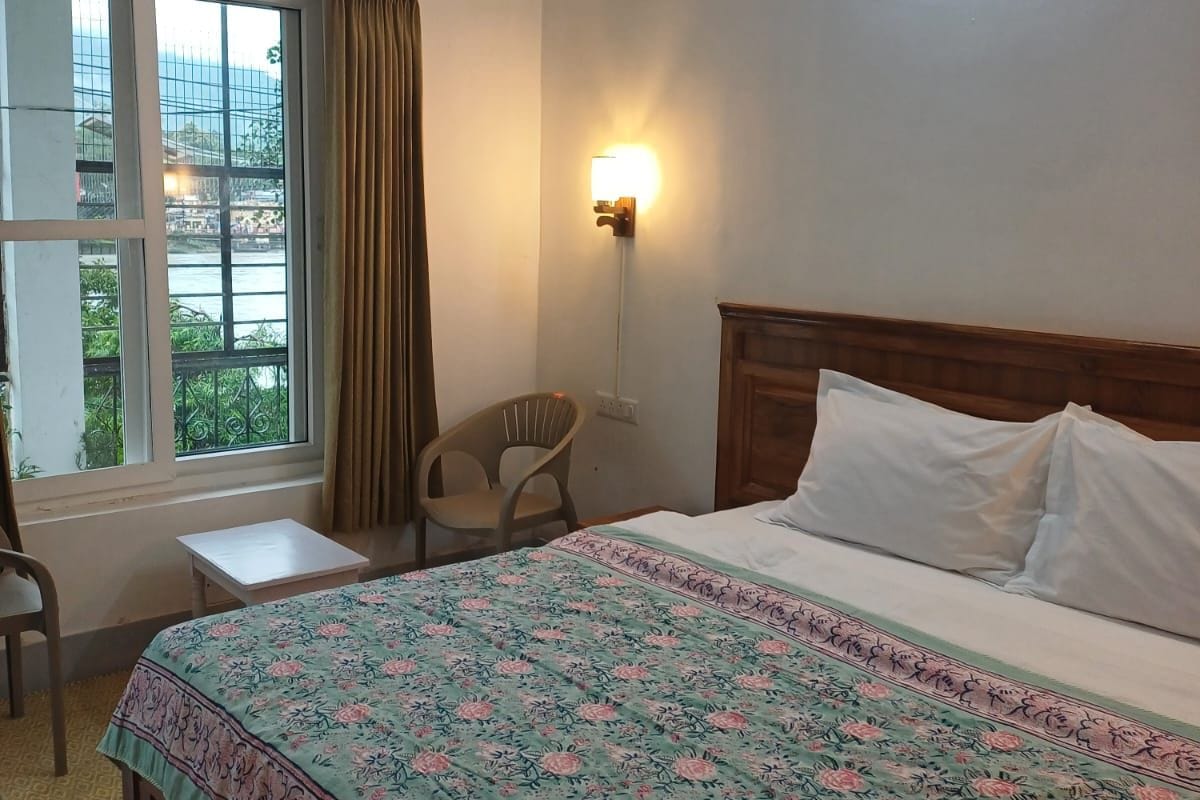 A hotel room with a bed, a chair, a small table, a window with curtains, and a wall-mounted light.