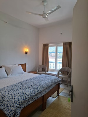 A hotel room with a bed, a ceiling fan, a window with curtains, and a view of the outside. - AJASRA-GANGA (Rishikesh)
