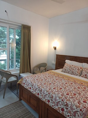 A bedroom with a bed, a chair, a desk, a window with curtains, and a wall-mounted light. - AJASRA-GANGA (Rishikesh)