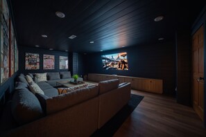 A modern living room with a sectional sofa, a coffee table, and a large TV screen displaying a movie scene. - Sleeps 36, Private Indoor Pool, Theater, Arcade, Hot Tub, 11 Mi to Dollywood (Sevierville)