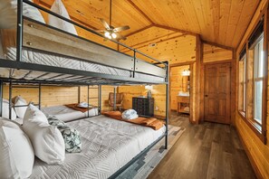 Quarto com beliche, ventilador de teto, paredes de madeira e banheiro visível por uma porta aberta.