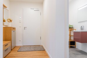 A hallway with a wooden floor, a white door, a small rug, a mirror, a vase with flowers, and a bathroom with a sink and a plant. - Yellow Duck - Beach Runner 4.4 | Sauna. Beach chair. Near the Baltic Sea. (Kellenhusen)