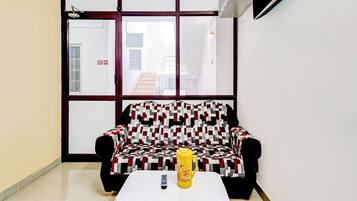 Una habitación con un sofá estampado, una mesita con un control remoto y una taza amarilla, y una ventana con letrero de salida de incendios.