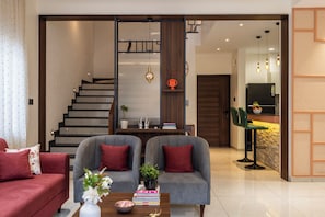 Una sala moderna con un conjunto de sofás, una mesa de centro con objetos decorativos, una estantería y una escalera que conduce a un nivel superior.