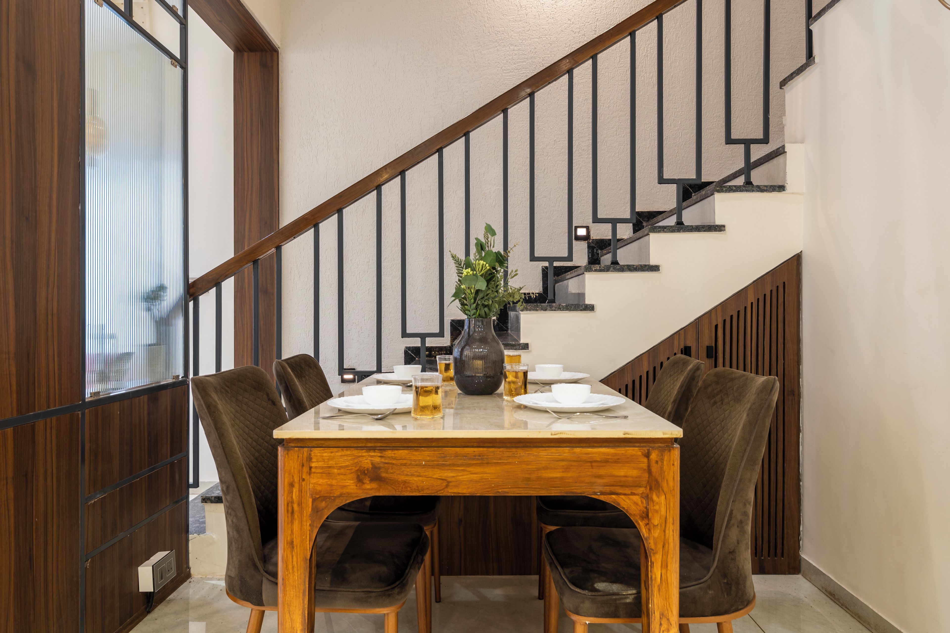 A dining area with a wooden table, four chairs, and a vase with flowers. A staircase with a wooden railing is visible in the background.