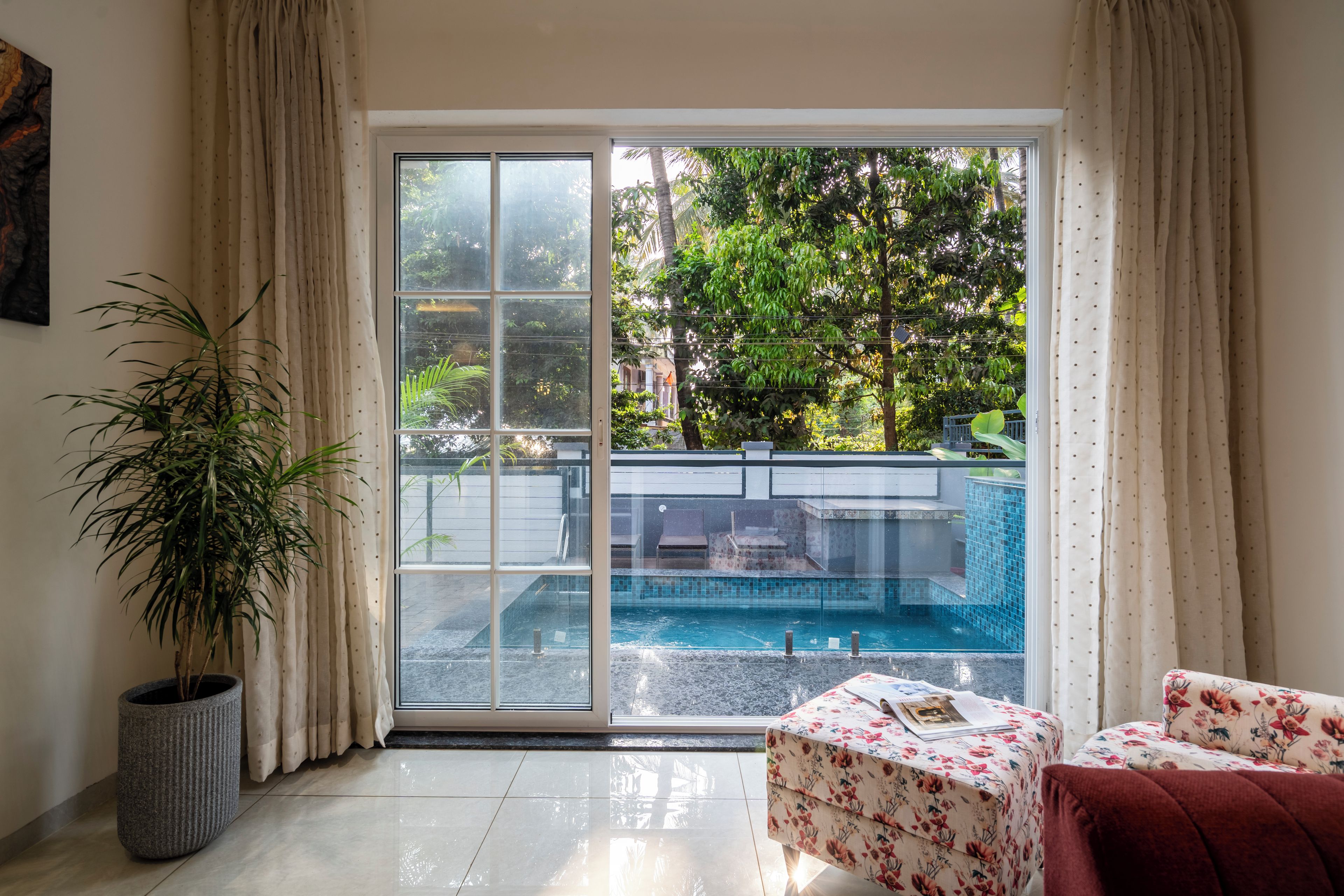 A room with a sliding glass door leading to a pool area, a potted plant, and a floral-patterned ottoman.