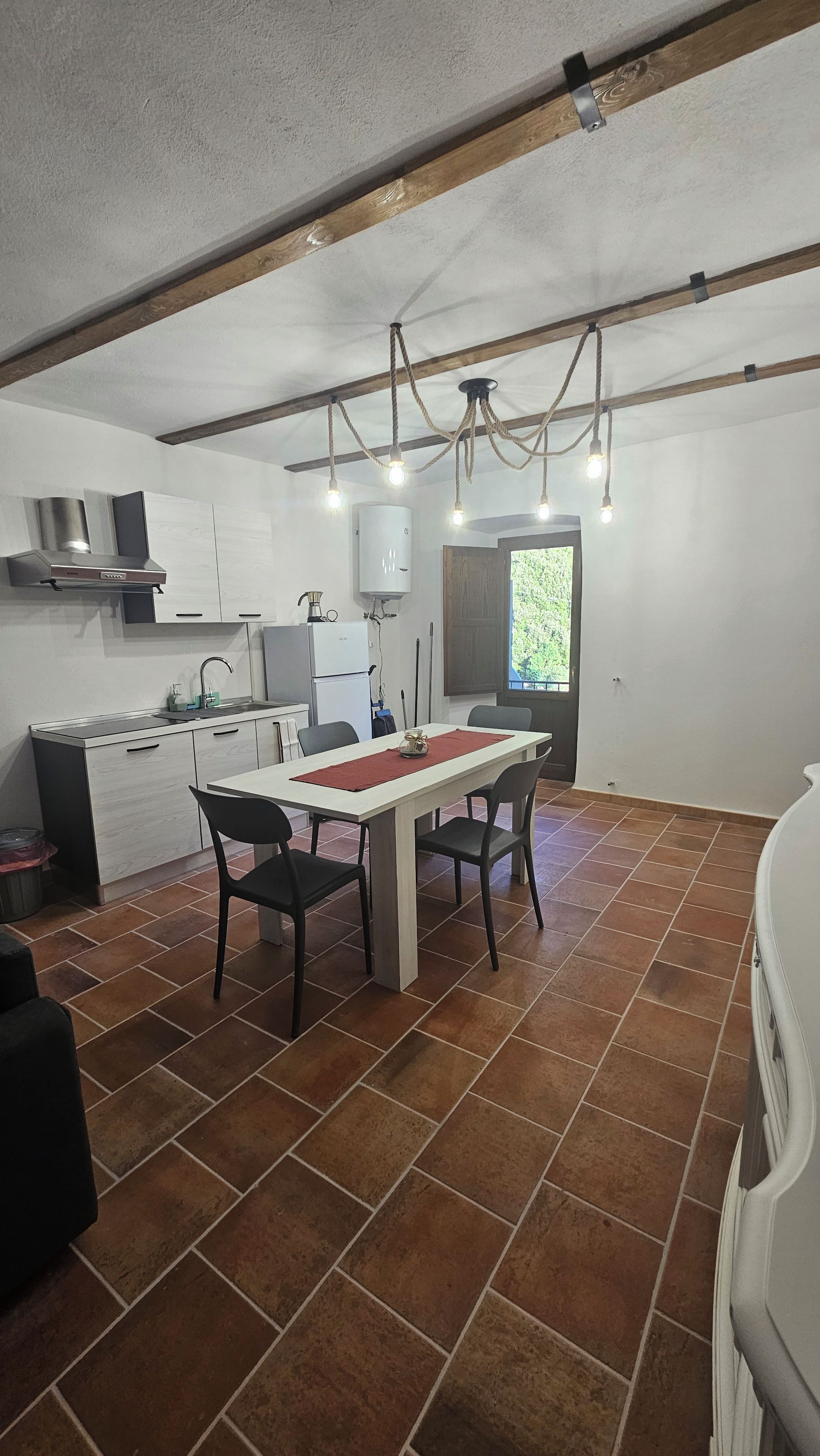 Cocina compacta con electrodomésticos modernos, comedor con camino de mesa rojo, y techo con vigas a la vista y luces colgantes.