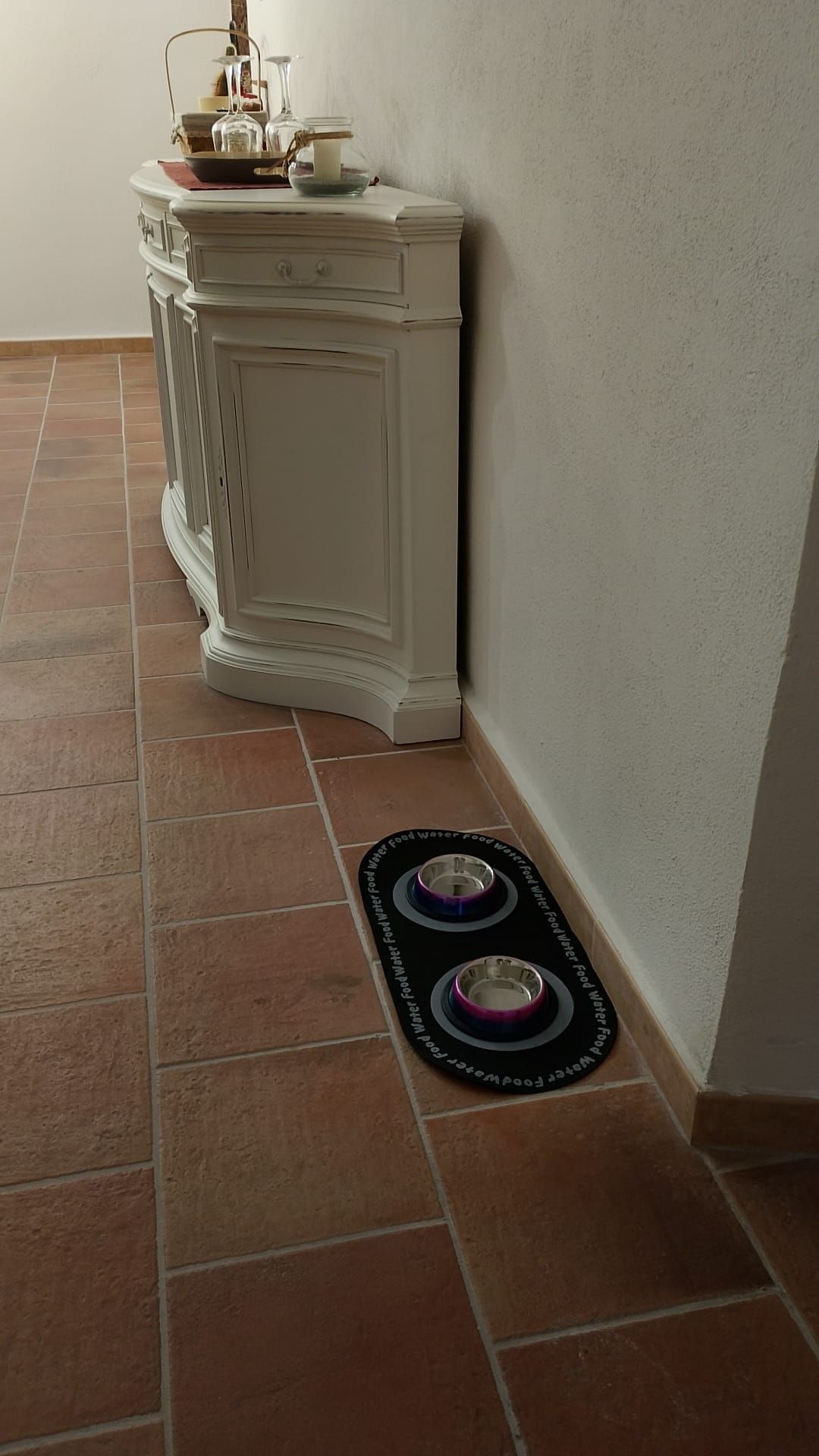 A corner of a room with a white cabinet against a wall, a tiled floor, and a pet water bowl on the floor.