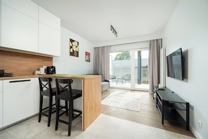 A modern kitchen with a wooden countertop, black bar stools, and a dining table. There is a sofa and a TV mounted on the wall. - RentPlanet - Apartamenty  Wagonowa (Warszawa)