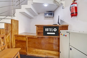 Un mostrador de recepción de hotel con detalles de madera, un extintor de incendios colgado en la pared y un sistema de intercomunicación de seguridad sobre el mostrador.