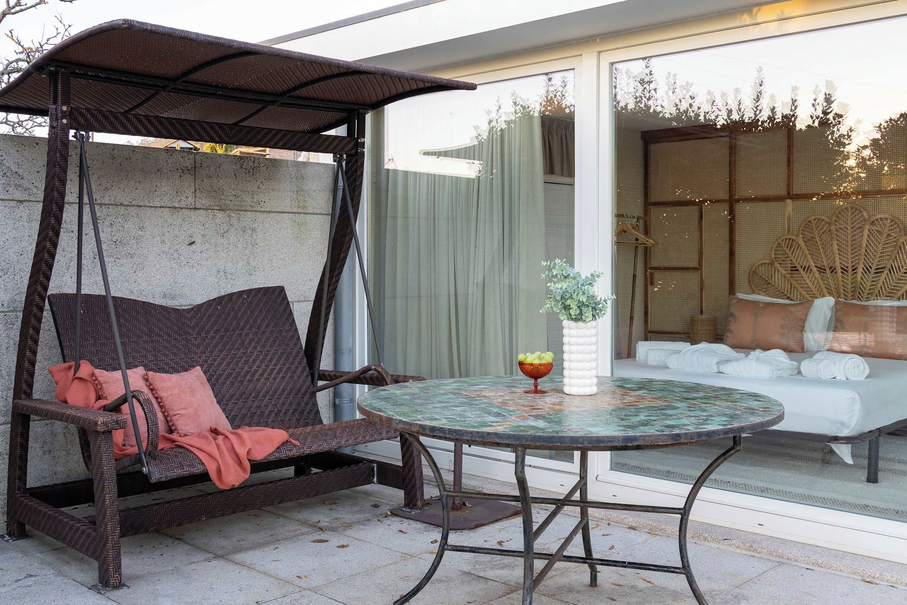 A wicker swing chair with cushions, a round table with a decorative bowl, and a sofa with cushions in a sunlit room.