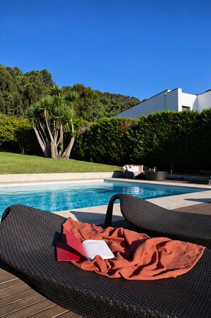 Una silla de descanso junto a la piscina, con una toalla y un libro, una piscina y un edificio blanco al fondo.