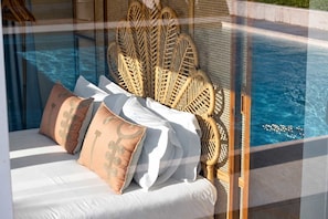 Cama con ropa blanca y almohadas decorativas, ubicada junto a una piscina y con cabecera de madera.