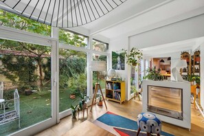 A modern living room with a large sliding glass door leading to a garden, a wooden floor, a round table, and a bookshelf. - Pilon | Ground-Floor Apartment with Private Garden in Montmartre (Paris)