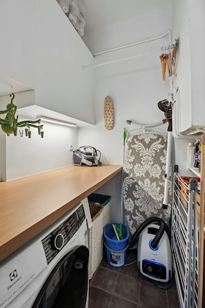 A laundry room with a washing machine, dryer, ironing board, and various cleaning tools. - Pilon | Ground-Floor Apartment with Private Garden in Montmartre (Paris)