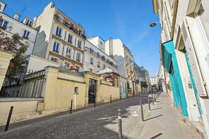 Une rue pavée bordée d’immeubles à plusieurs étages, dont certains sont dotés de balcons et de volets.