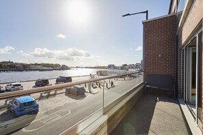 A balcony with a view of a waterfront, a parking area with cars, and a building with a brick facade. - Host & Stay | Fish Quay View (Tynemouth)