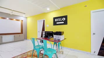 Habitación de hotel con pared amarilla, un letrero que indica que es un hotel de servicios corporativos, un escritorio con una impresora, dos sillas turquesa y una alfombra estampada.
