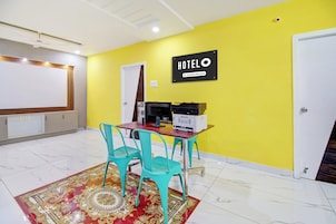 Habitación de hotel con pared amarilla, un letrero que indica que es un hotel de servicios corporativos, un escritorio con una impresora, dos sillas turquesa y una alfombra estampada.