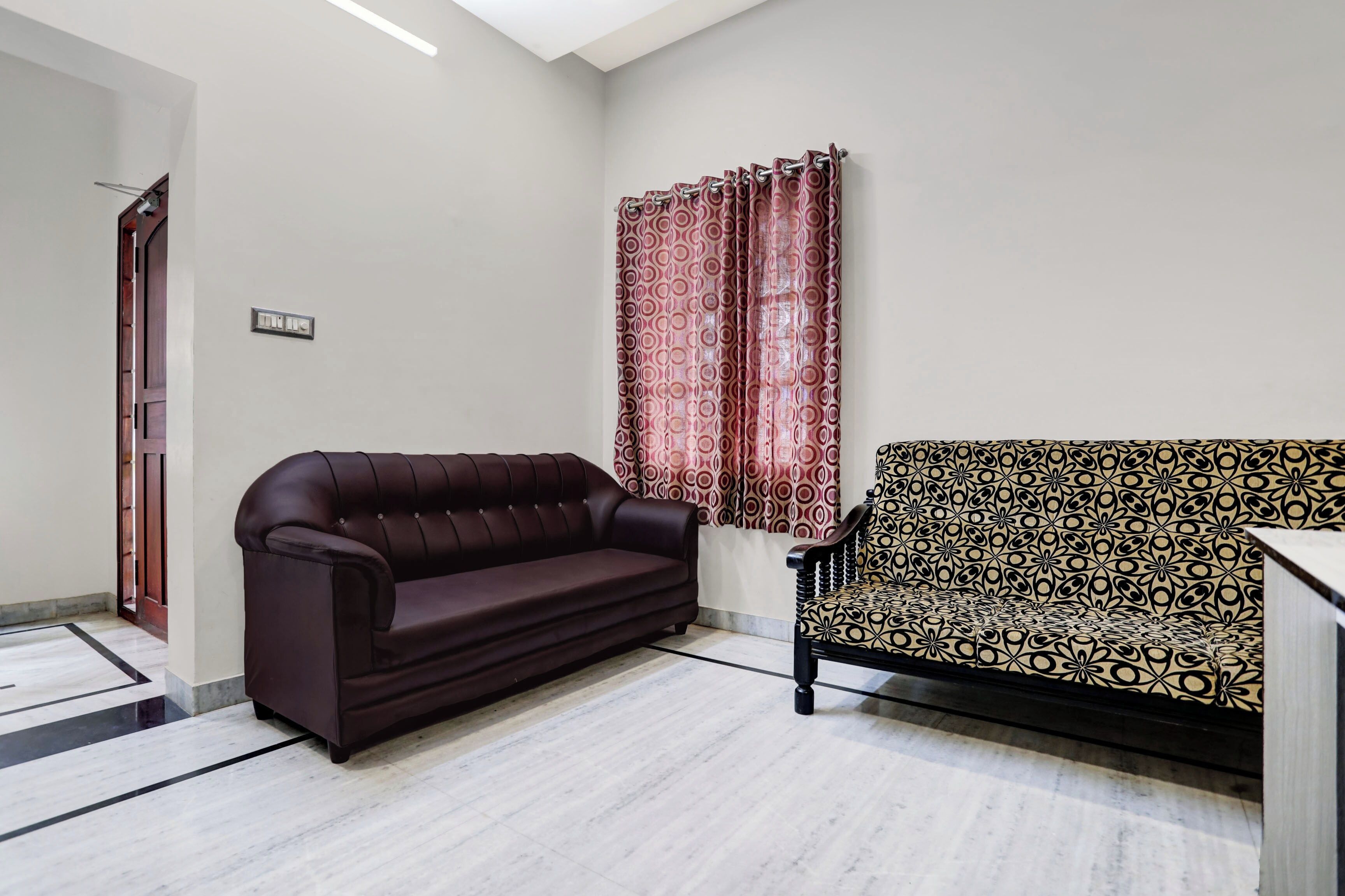 A room with a dark brown sofa, a patterned armchair, and a window with red curtains.