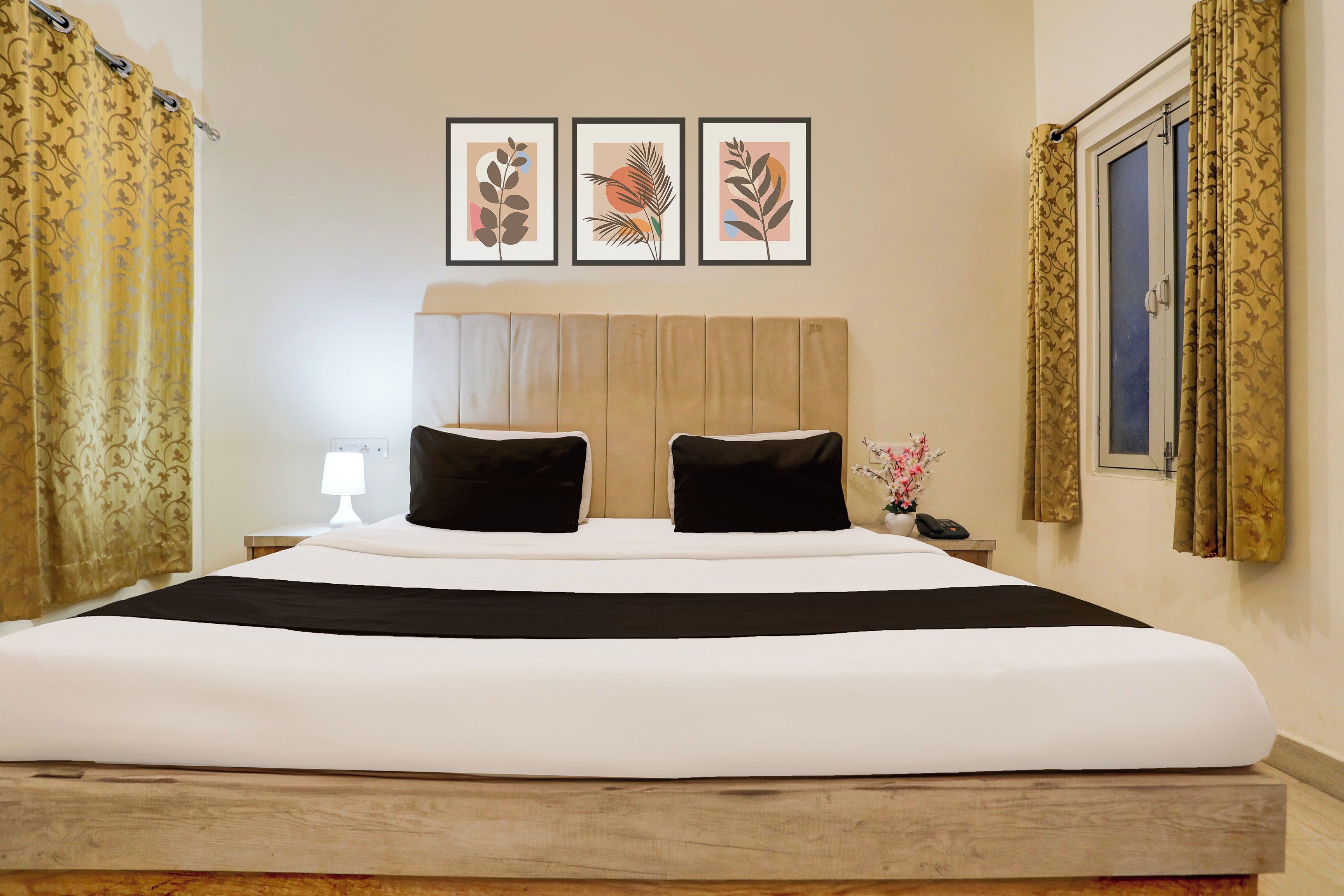 Habitación de hotel con una cama grande, dos almohadas negras, un cabecero de madera, una mesita de noche con una lámpara, un teléfono y tres láminas botánicas enmarcadas en la pared.