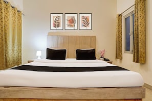 Ein Hotelzimmer mit einem großen Bett, zwei schwarzen Kissen, einem hölzernen Kopfteil, einem Nachttisch mit Lampe, einem Telefon und drei gerahmten Pflanzenbildern an der Wand.