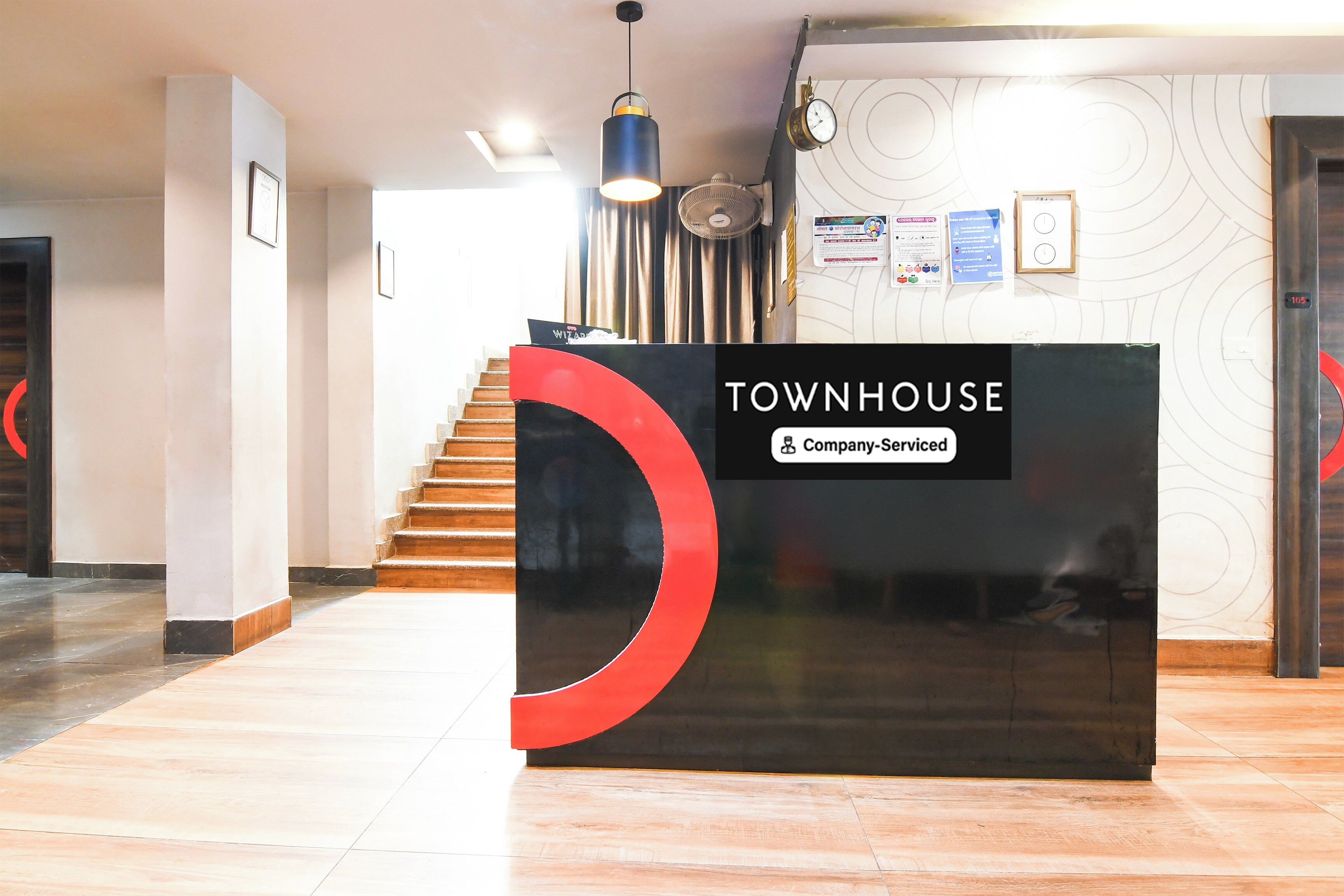 Un mostrador de recepción con un letrero que dice 'TOWNHOUSE' y 'Company Serviced' en un vestíbulo moderno con una escalera y un revestimiento de pared con diseño.