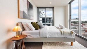 Un dormitorio moderno con una cama grande, una lámpara de noche, vistas de la ciudad a través de un amplio ventanal y un cuadro en la pared.