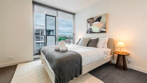 A modern bedroom with a large bed, a bedside table with a lamp, a window with a view of buildings, and a painting on the wall. - Richmond Rhythm | City Living & Lifestyle (Richmond)
