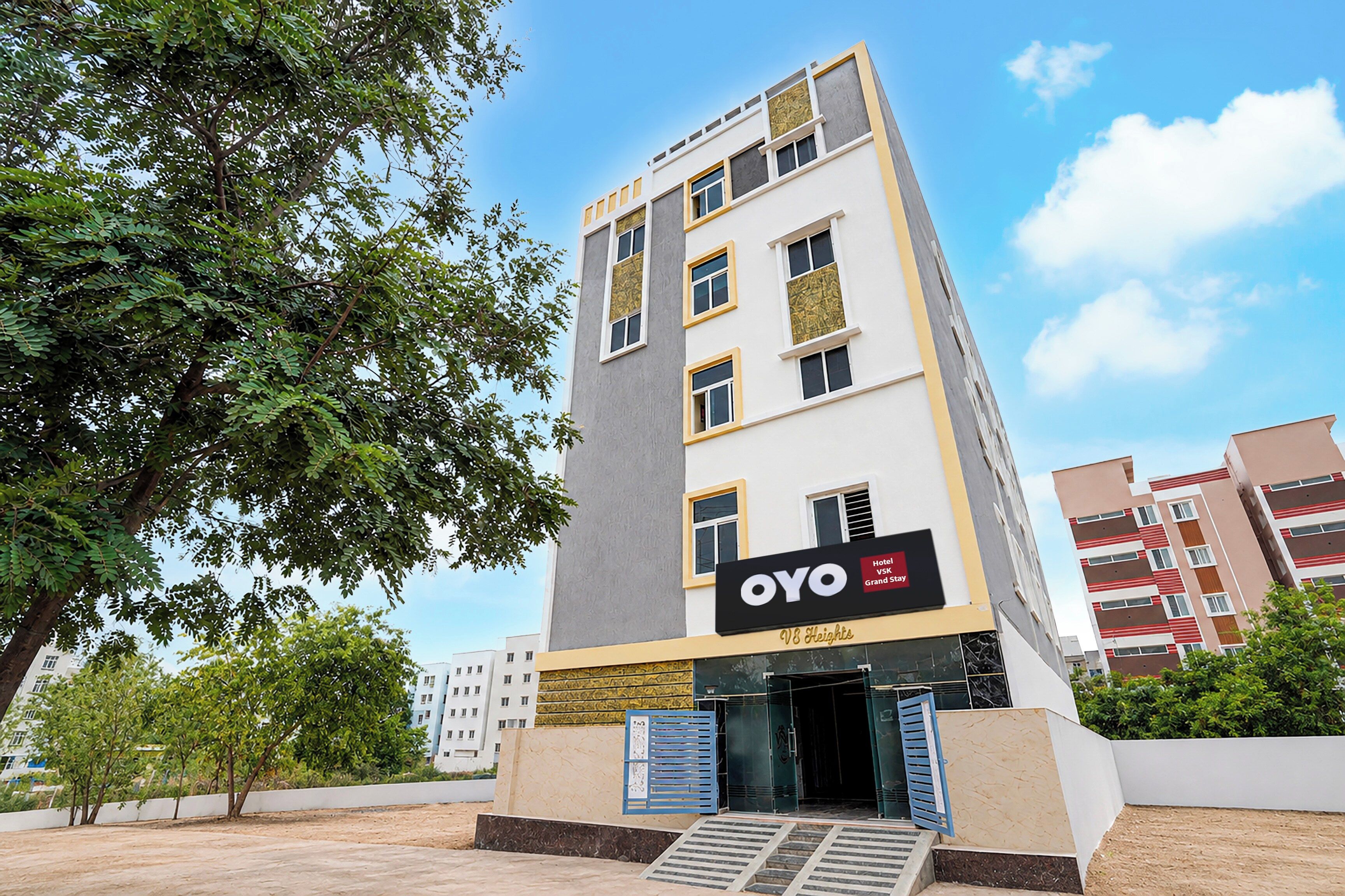 A modern hotel building with a prominent sign for 'OYO VSK Grand Stay' and 'V8 Heights' on the facade.