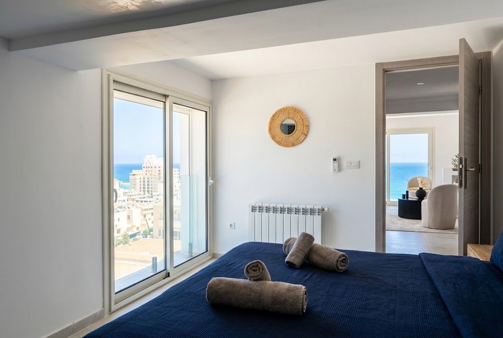 A bedroom with a large window, a bed with rolled towels, a radiator, and a view of the city and sea.