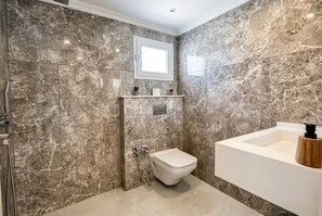 Ein Badezimmer mit Marmorwand, Toilette in Weiß, Dusche und Waschbecken in Weiß mit Holz-Seifenschale.
