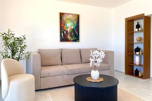 A living room with a beige sofa, a round coffee table with a white vase, a painting on the wall, and a wooden bookshelf. - sandra 18 (Tunis)