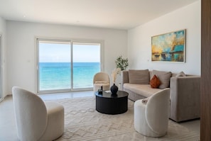Ein modernes Wohnzimmer mit einer großen Schiebeglastür, die auf einen Balkon führt, einem beigen Sofa, zwei Sesseln und einem schwarzen Couchtisch.