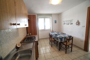 A kitchen with a gas stove, a sink, wooden cabinets, a dining table with chairs, and a window with blinds. - Apartment 'Kambusa Homes - Al Porto - Destra' with Sea View, Wi-Fi and Air Conditioning (Torre Vado)