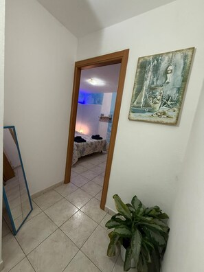 Ein Flur mit Fliesenboden führt zu einem Schlafzimmer mit Bett und Nachttisch. An der Wand befinden sich ein Blumentopf und ein gerahmtes Bild.