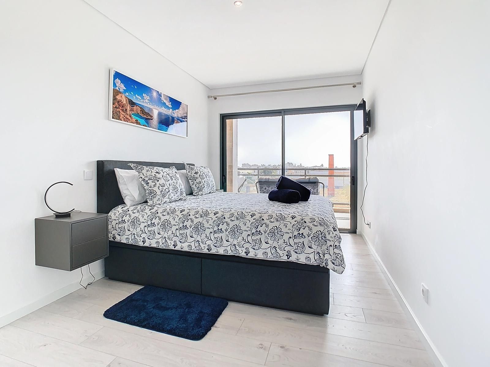 Ein modernes Schlafzimmer mit einem großen Bett, einem Nachttisch, einem blauen Teppich und Blick auf die Stadt durch ein großes Fenster.