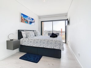 Ein modernes Schlafzimmer mit einem großen Bett, einem Nachttisch, einem blauen Teppich und Blick auf die Stadt durch ein großes Fenster.