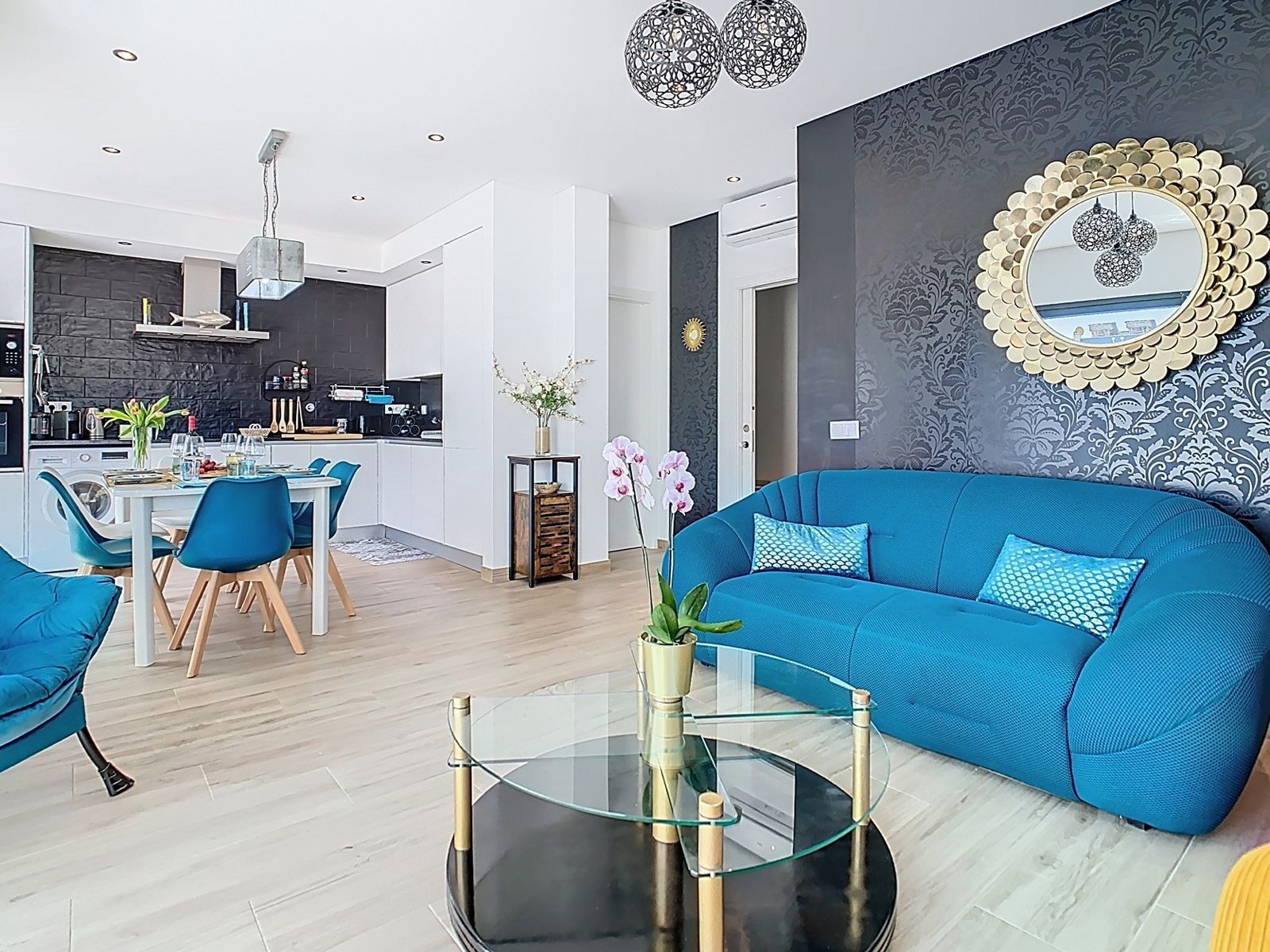 A modern living room with a blue sofa, a glass coffee table, and a kitchen in the background.