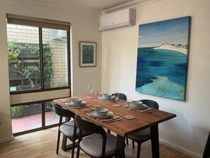 Un coin repas avec une table dressée pour quatre personnes, un tableau représentant une scène de plage et une fenêtre donnant sur un paysage verdoyant.