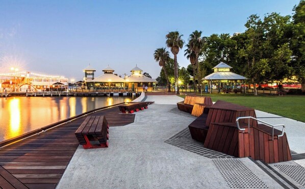 Une terrasse au bord de l’eau, dotée de mobilier en bois, un quai et une rangée de bâtiments coiffés de toits en dôme.
