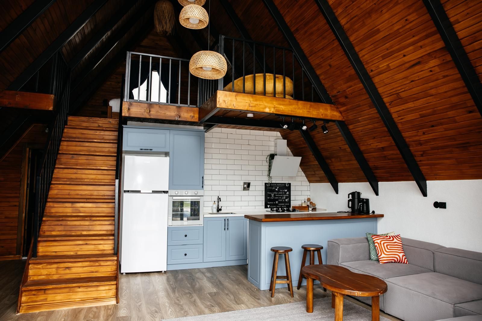 A loft-style kitchen with a wooden staircase, a dining area with a sofa and stools, and a hanging light fixture.