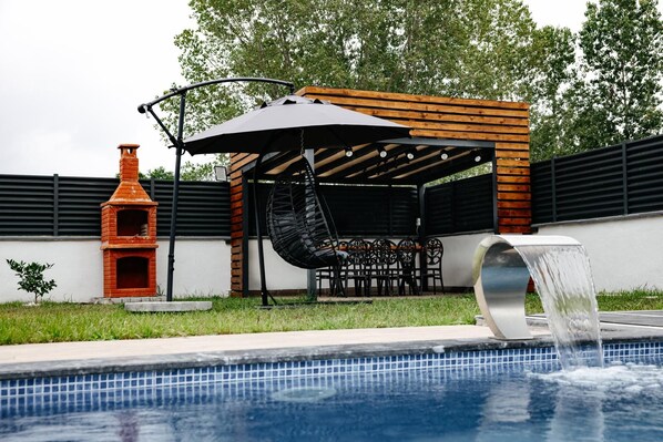 A swimming pool with a water feature, a brick oven, and a covered seating area with chairs and tables.