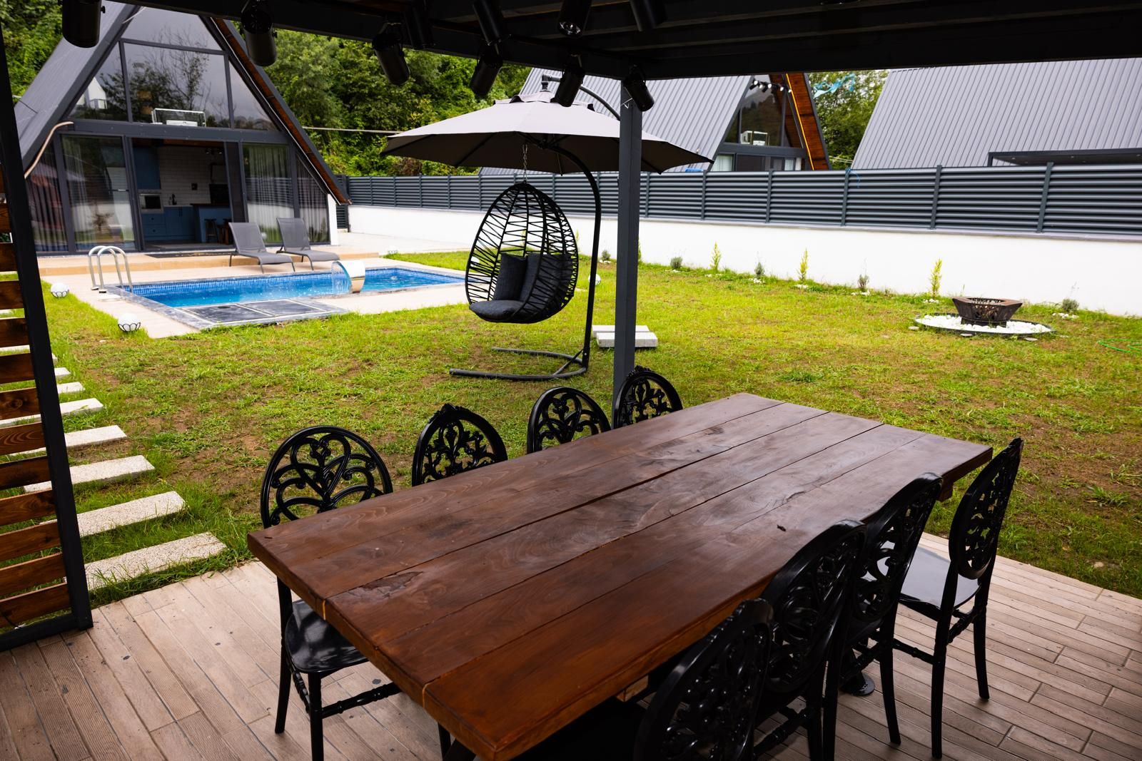 A wooden table with chairs, a hanging chair, a pool, and a building in the background.