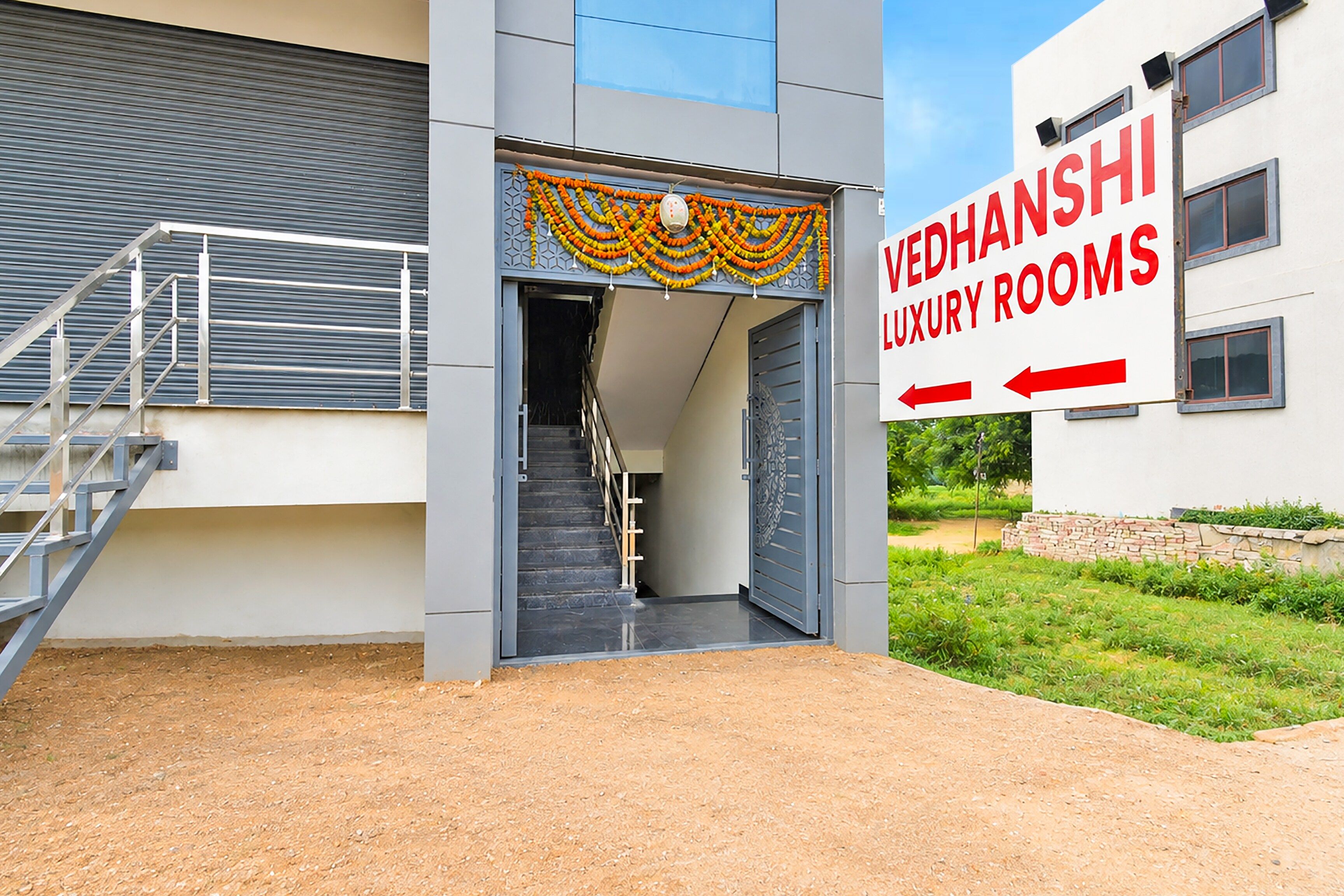 Una entrada de edificio con una escalera que lleva a una puerta, un letrero que indica 'VEDHANSHI LUXURY ROOMS' y un camino de acceso con portones.