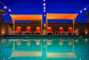 Piscina amb gandules sota una zona d'ombra, envoltada per una tanca i il·luminada per llums penjants.