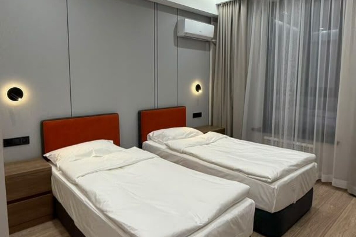 Una habitación de hotel moderna con dos camas individuales, piso de madera, un ventanal con cortinas traslúcidas y aire acondicionado instalado en la pared.