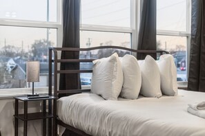 A hotel room with a bed, a nightstand, a lamp, and a view of the outside. - 6BR/4BA Duplex | 10 Minutes to Downtown, Lucas Oil & Gainbridge (Indianapolis)