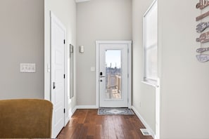 A hallway with a white door leading to a balcony, a brown chair, and a wall-mounted light switch. - 6BR/4BA Duplex | 10 Minutes to Downtown, Lucas Oil & Gainbridge (Indianapolis)
