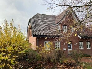 A brick building with a tiled roof, surrounded by deciduous trees and shrubs. - Strandstrasse 28, Half of the House on the Left (Nieblum)